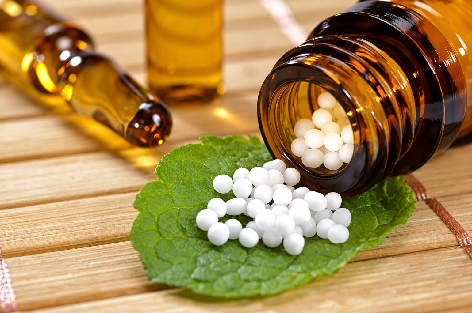 Des granules blancs d’homéopathie s’échappent d’un flacon en verre brun couché sur une feuille verte, posée sur un support en bois clair.