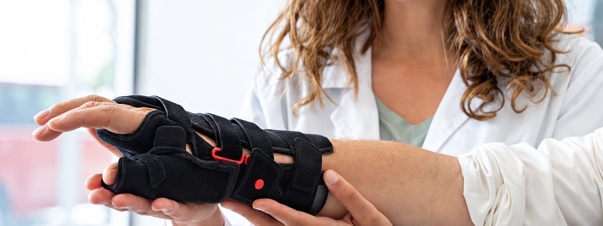 Une professionnelle de santé en blouse blanche ajuste une orthèse de poignet noire à sangles sur la main et l’avant-bras d’un patient.